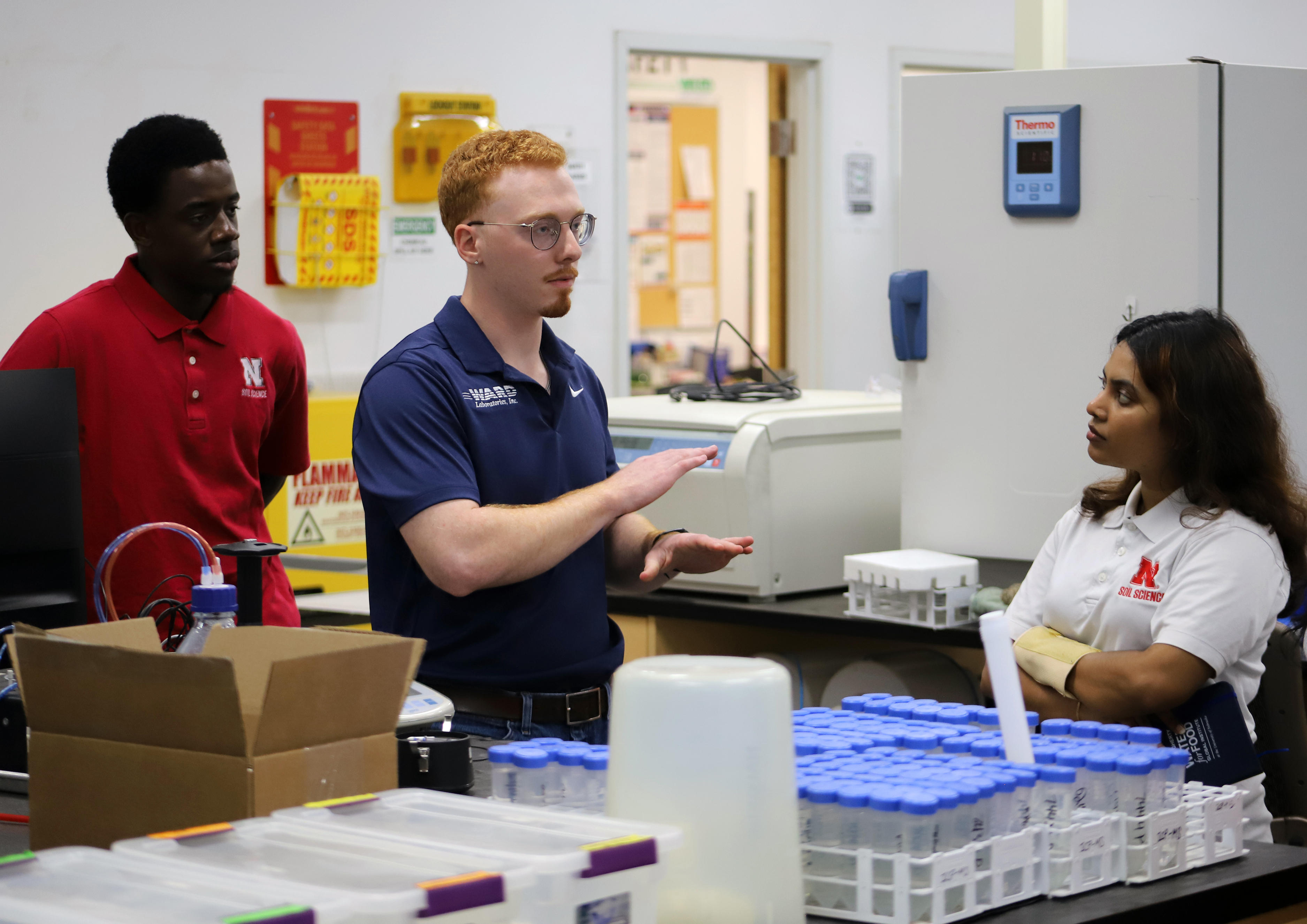 Three people converse in a lab surrounded by equipment and supplies.