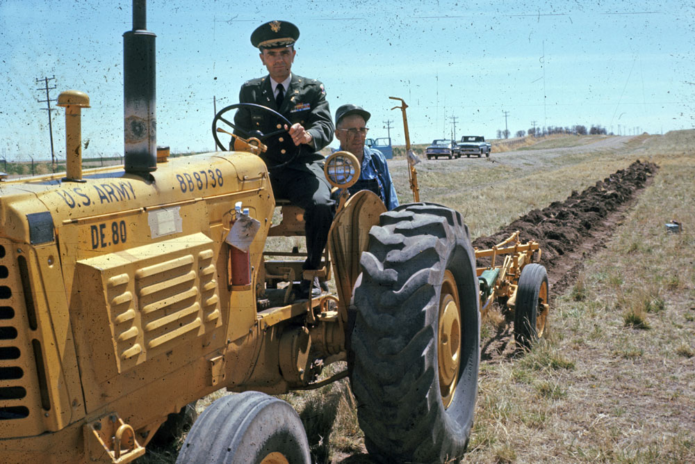 military man on tractor with a farmer behind him.