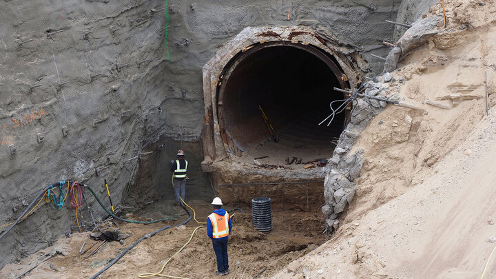 Large tunnel under construction in a sandy excavation site, with workers wearing safety gear nearby.