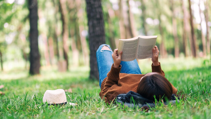 Person lying on grass, reading a book, with a straw hat nearby in a sunny park.