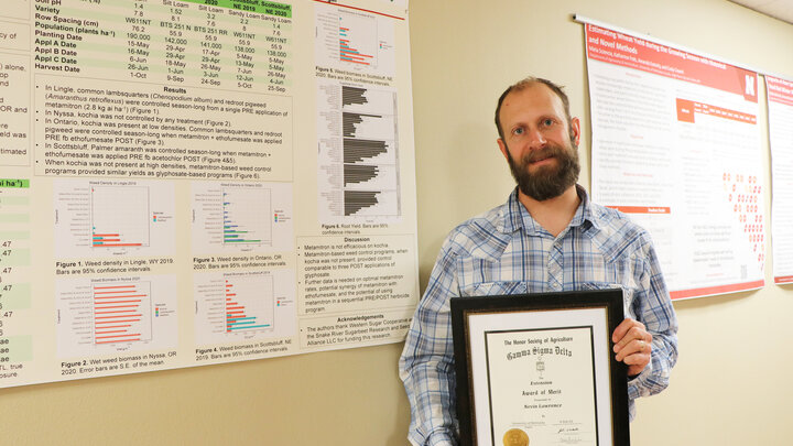 Man holding framed certificate in front of scientific posters on a beige wall.