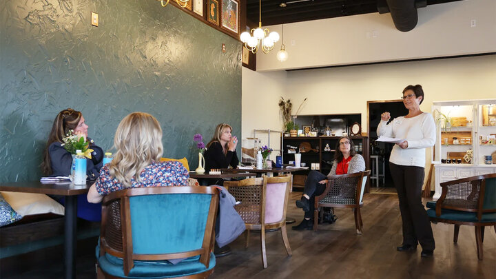 several women sit at tables in a cafe listening to woman speaker.