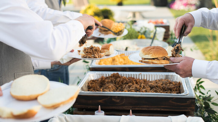 People serving themselves pulled pork and sides at an outdoor buffet table.