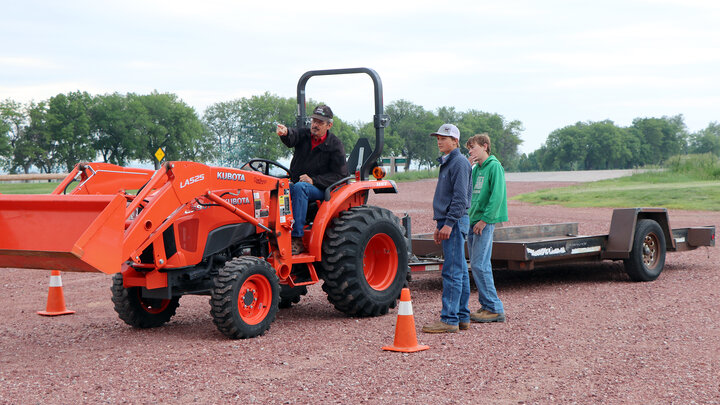 Man driving orange tractor while gesturing to two people standing near a trailer outdoors.